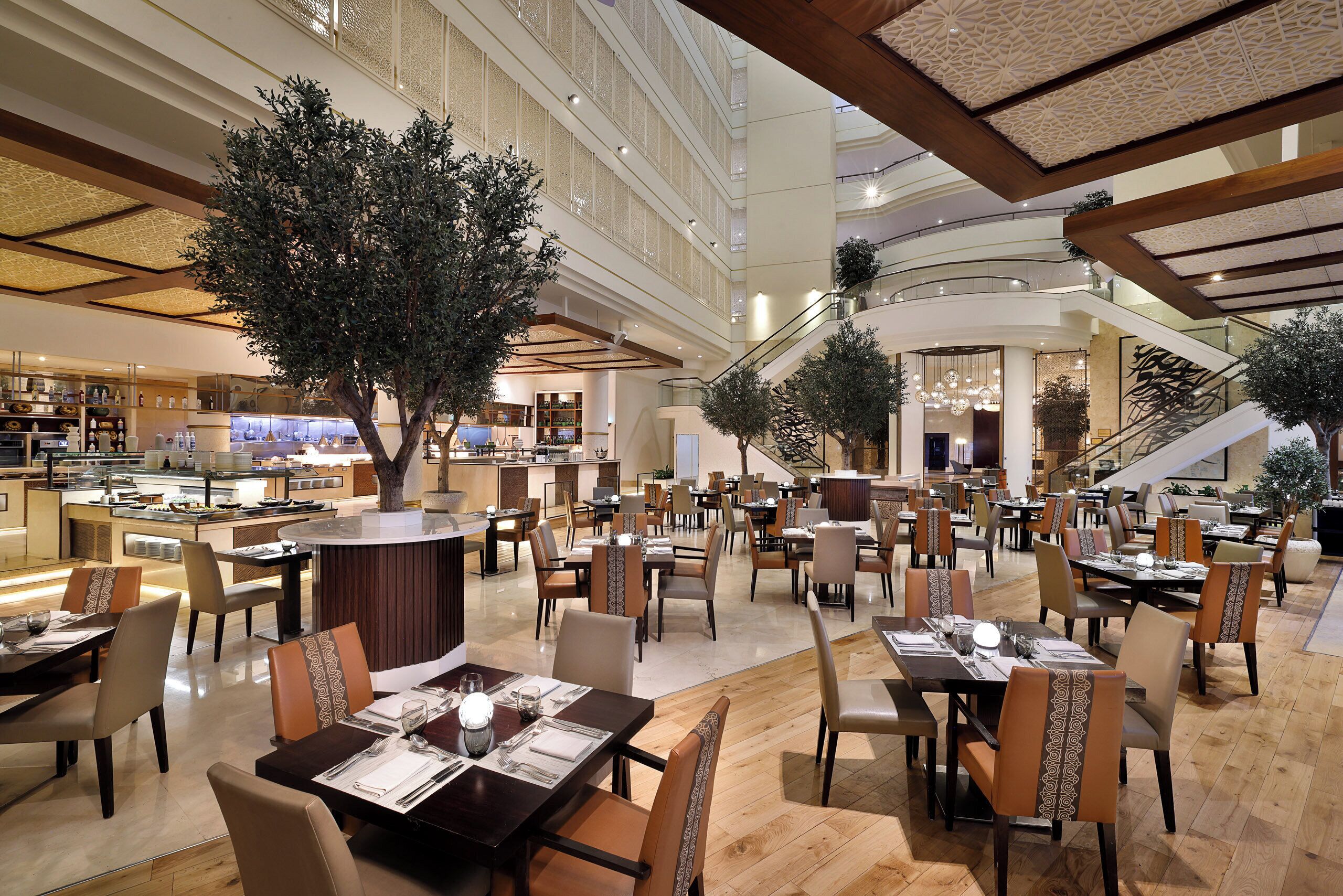 A spacious hotel lobby with multiple dining areas, a central atrium with a large tree, and a staircase leading to upper floors.