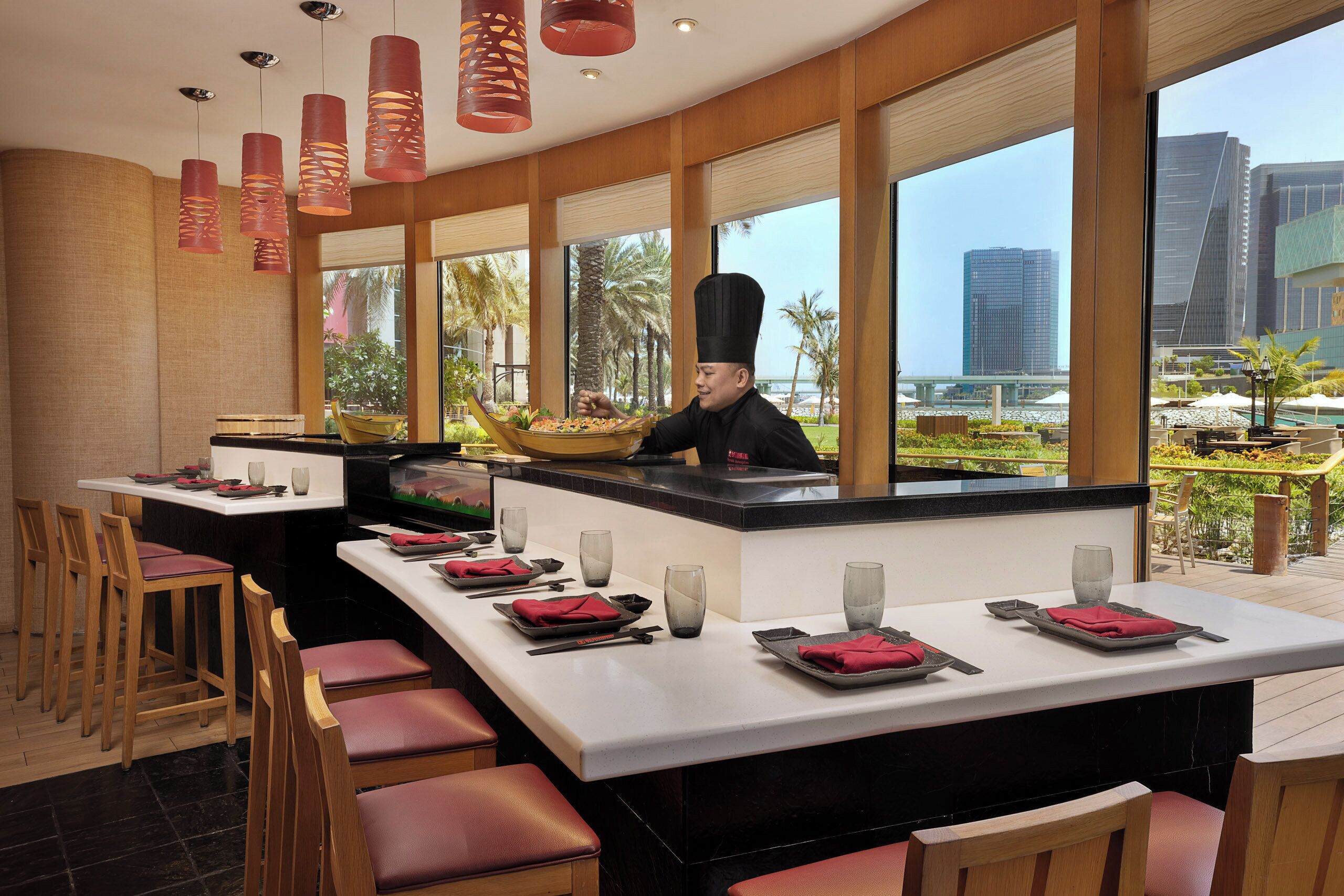 A restaurant with a chef preparing food, large windows with a view of palm trees and buildings, and neatly set dining tables with red napkins and glasses.