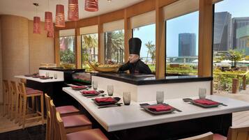 A restaurant with a chef preparing food, large windows with a view of palm trees and buildings, and neatly set dining tables with red napkins and glasses.