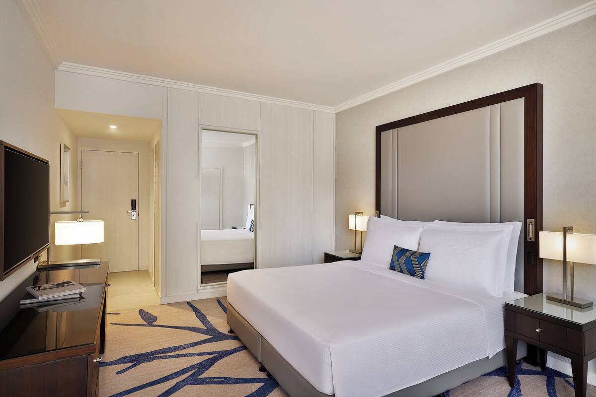 A hotel room with a large bed, bedside tables with lamps, a TV, a mirror, and a door leading to another room.