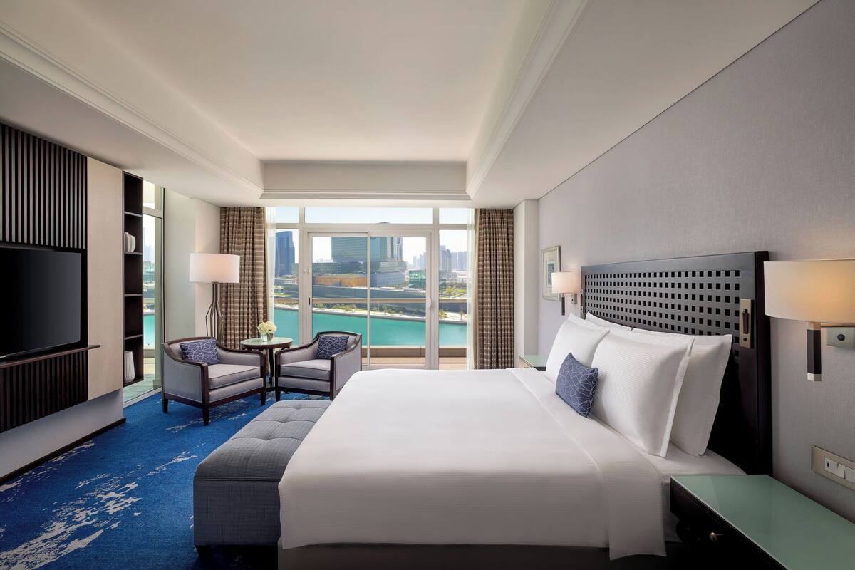 A hotel room with a large bed, a TV, a seating area with a table and chairs, and a view of a cityscape through large windows.