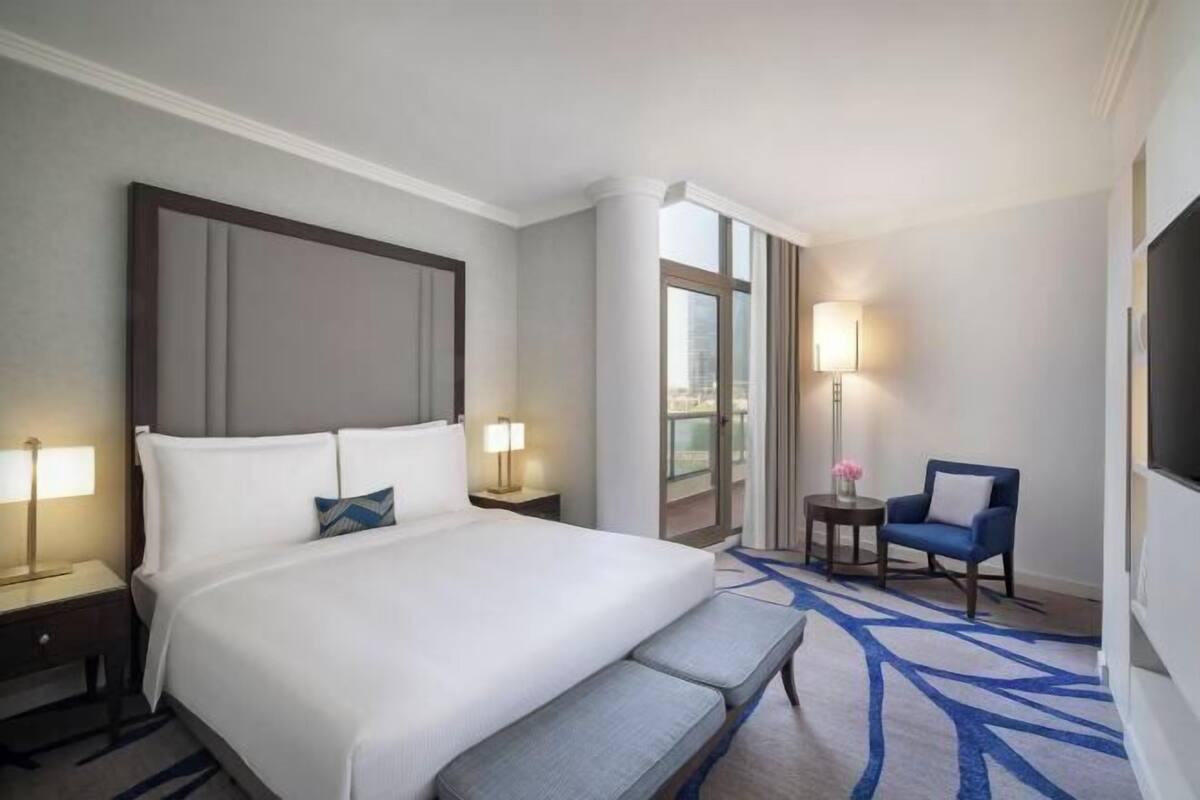 A hotel room with a large bed, bedside tables with lamps, a bench, a chair, and a TV.