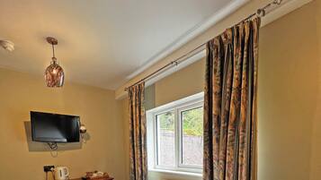A hotel room with a bed, a television mounted on the wall, a radiator, a window with curtains, and a table with a kettle and a tray.