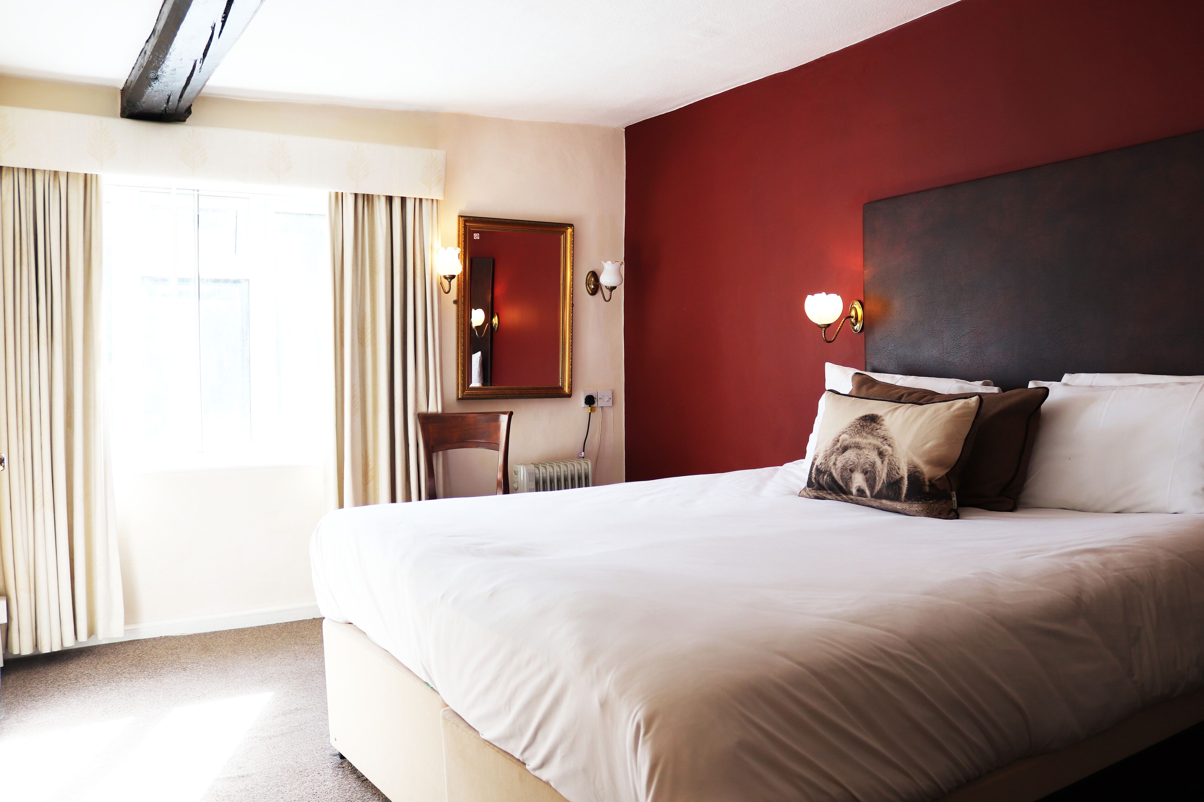 A hotel room with a large bed, a red accent wall, a mirror, and a window with curtains.