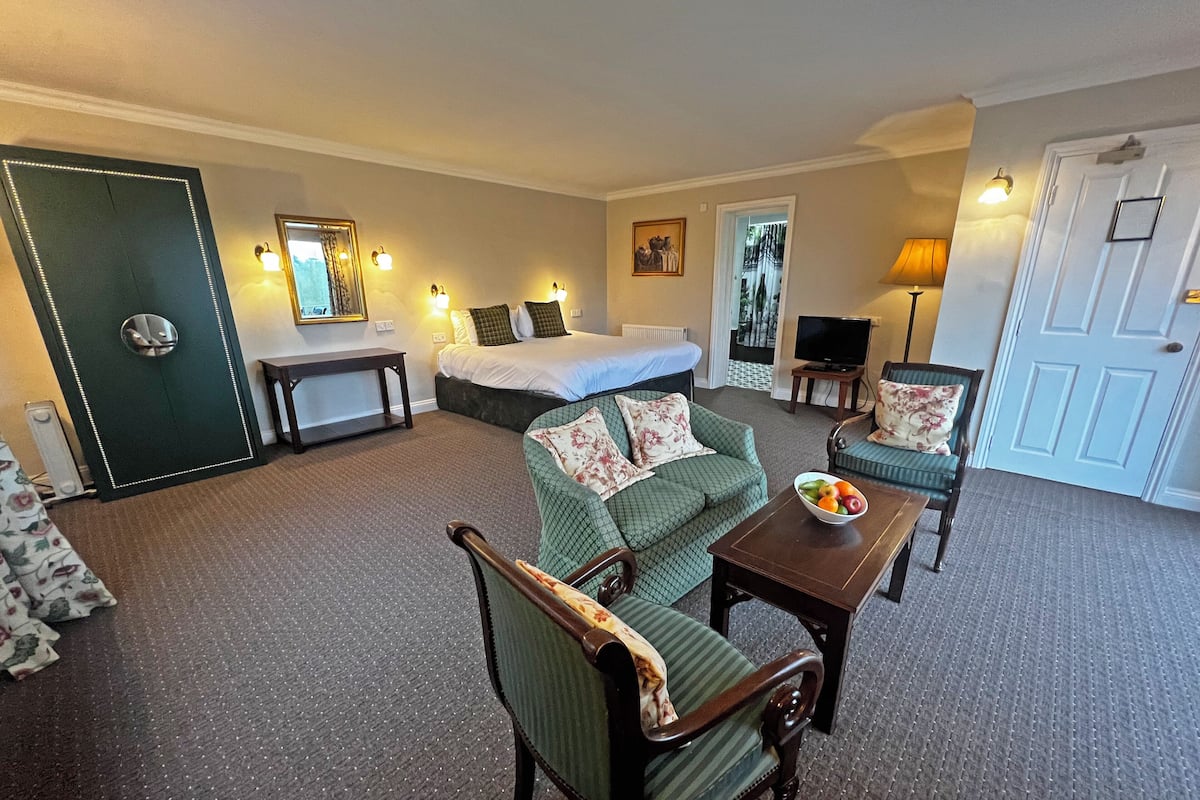 A hotel room with a bed, a sofa, a dining table with a fruit bowl, a TV, and a door.