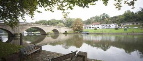 Une rivière paisible avec un pont de pierre, un banc et une zone herbeuse, avec un bâtiment en arrière-plan.