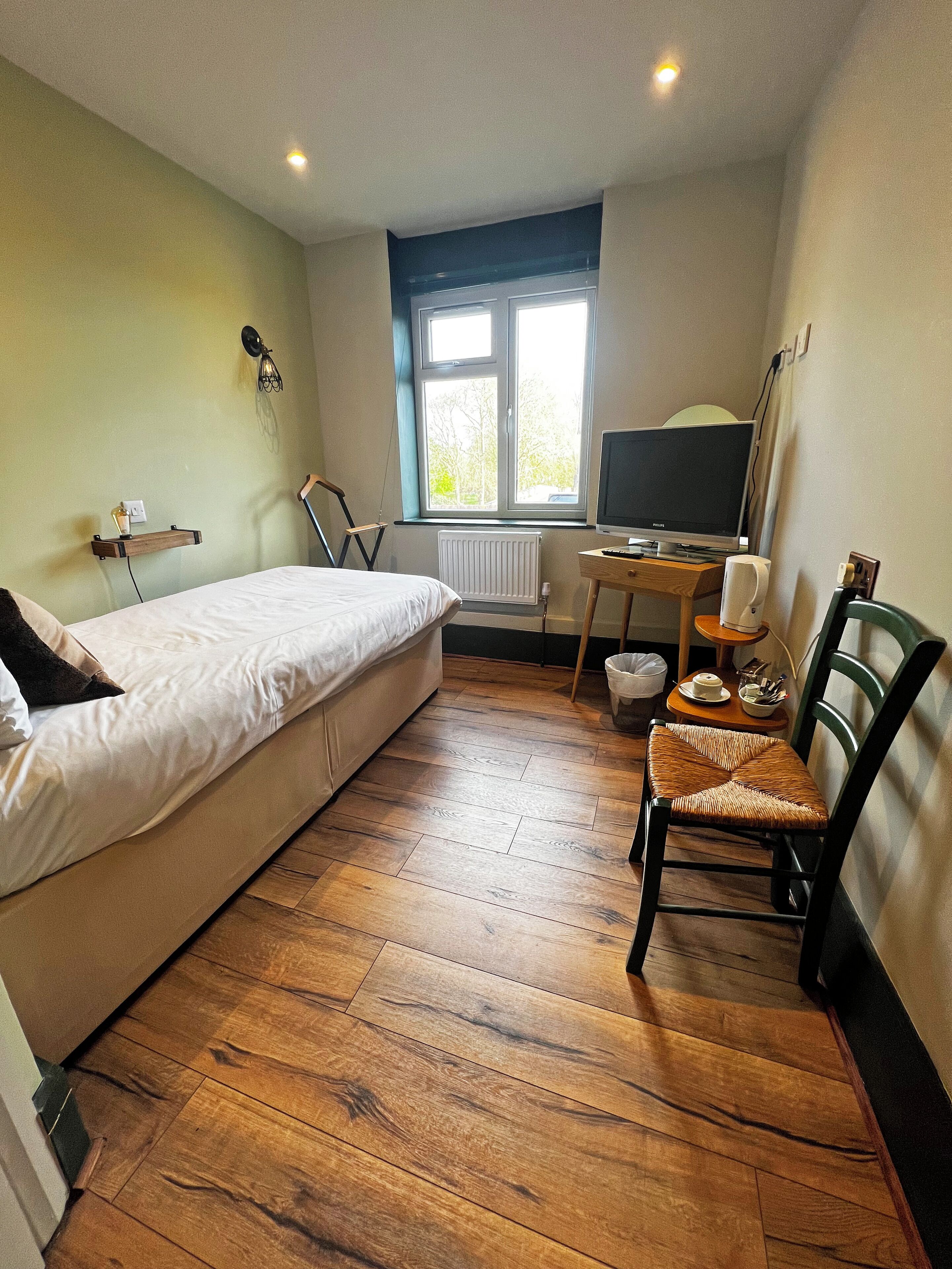 A bedroom with a bed, a desk with a computer, a chair, and a window with a view of trees.