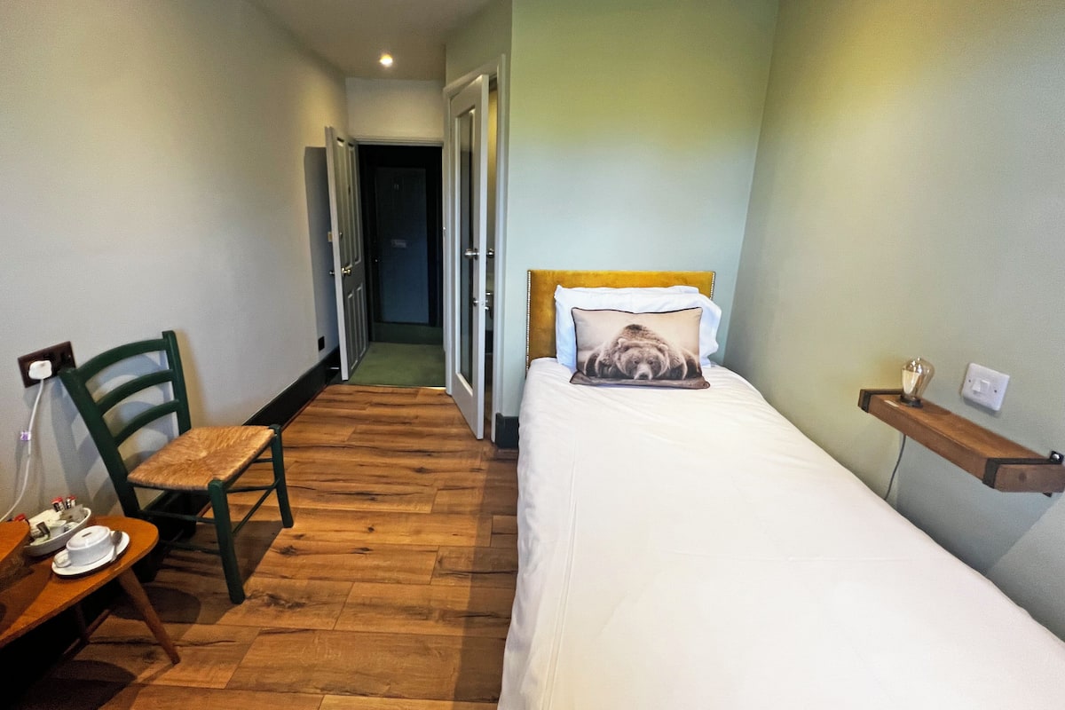 A small, cozy room with a wooden floor, a single bed with a pillow, a chair, and a small table with a cup and saucer.