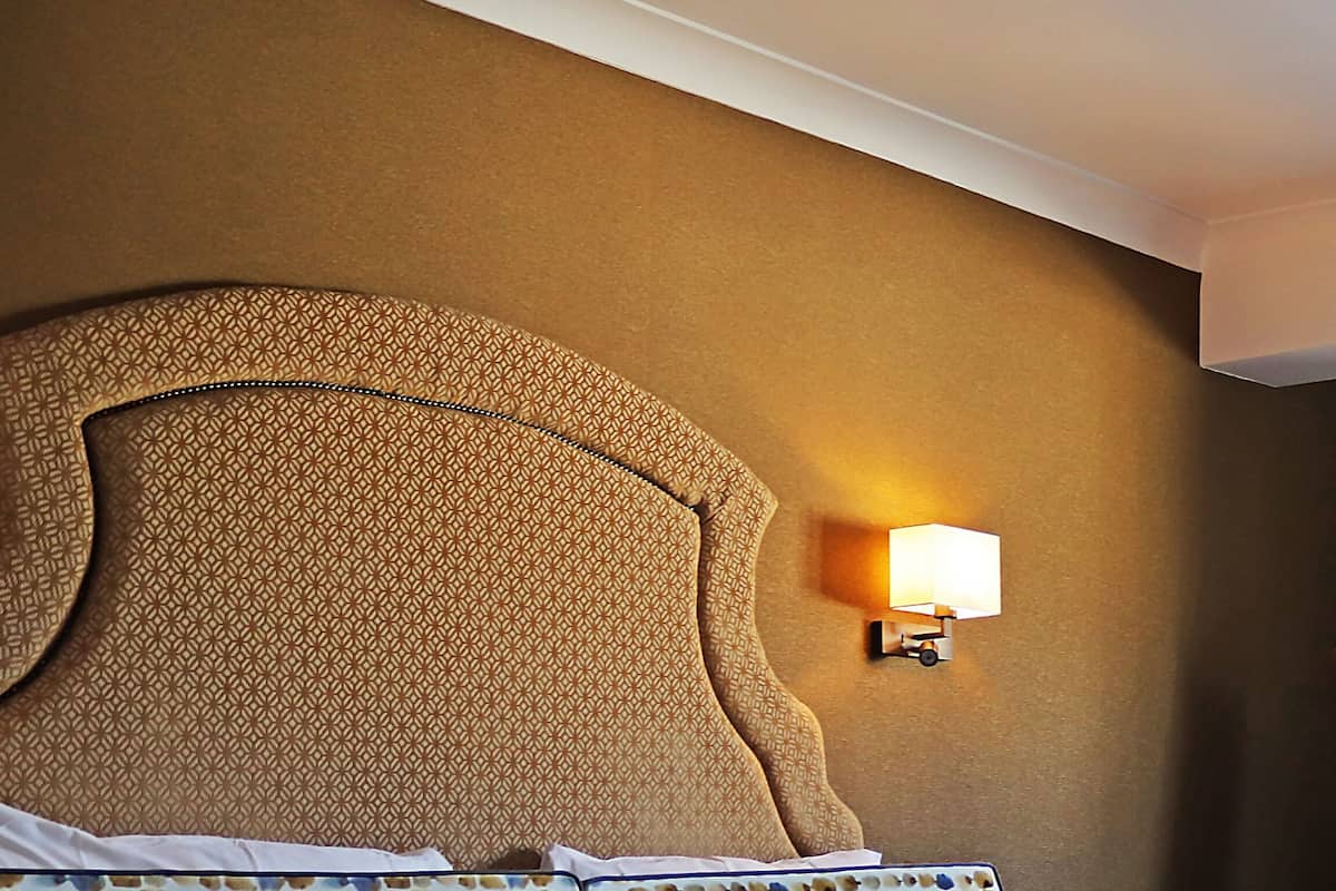 A hotel room with a large, textured headboard, a wall-mounted lamp, and patterned pillows.
