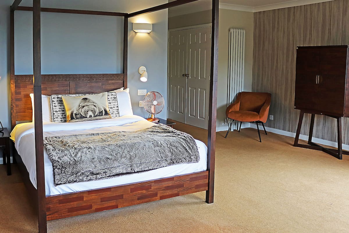 A four-poster bed with a wooden headboard, a bedside table, a wall-mounted fan, and a chair in a room with a door and a radiator.