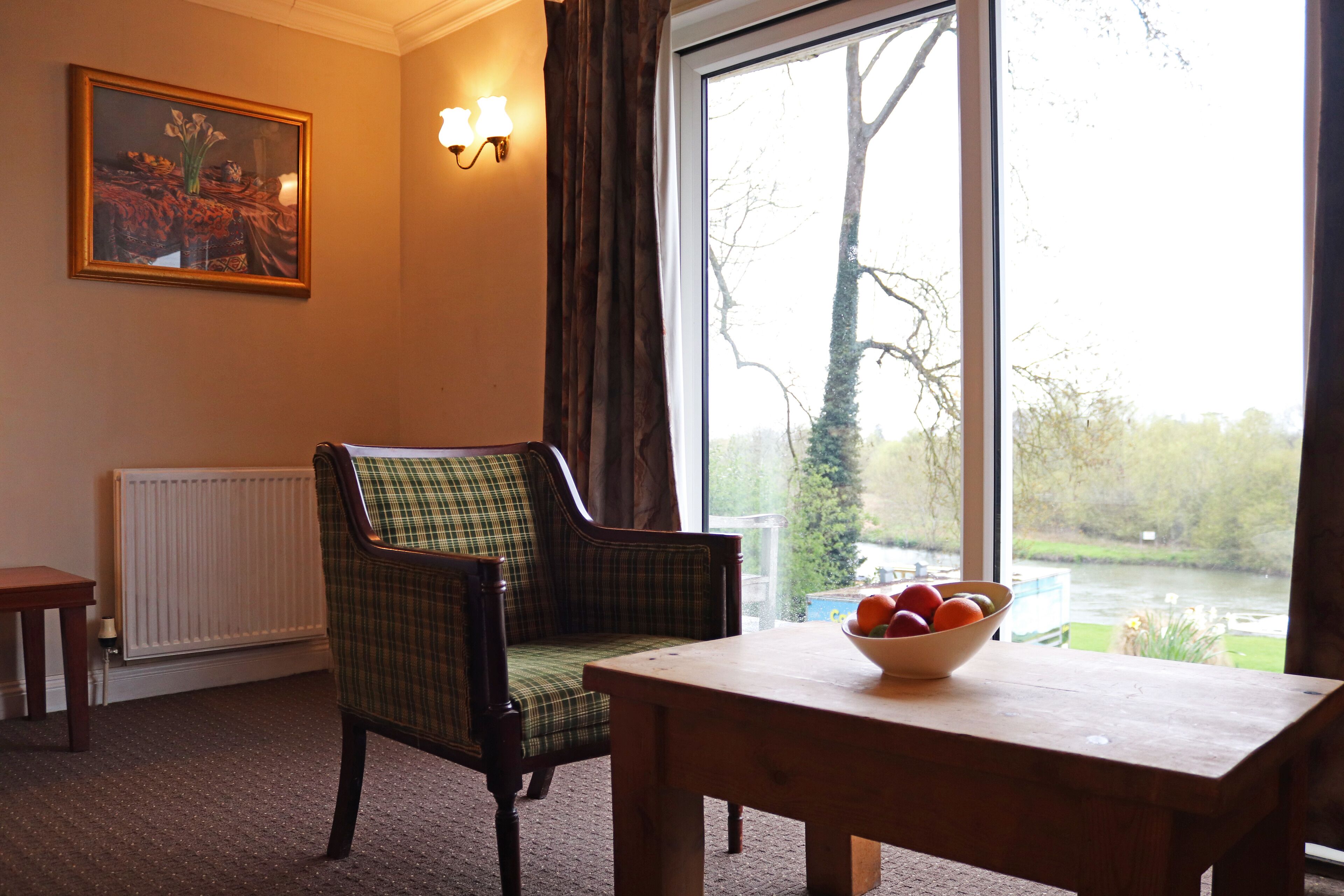 A room with a view of a river, a plaid chair, a wooden table with a bowl of fruit, a radiator, and a painting on the wall.