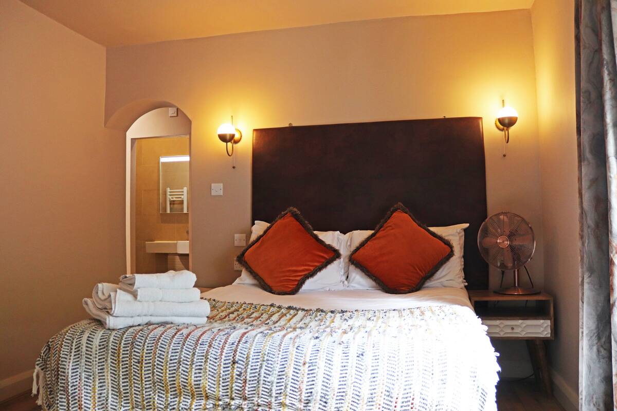 A hotel room with a large bed, two wall-mounted reading lights, a bedside table with a fan, and a mirror in the corner.