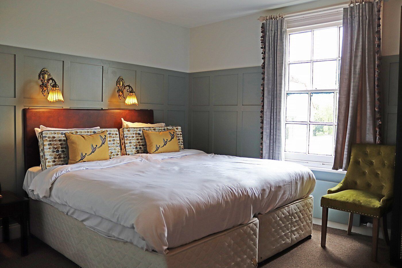 A bedroom with a large bed, two wall-mounted lamps, a window with curtains, and a green armchair.
