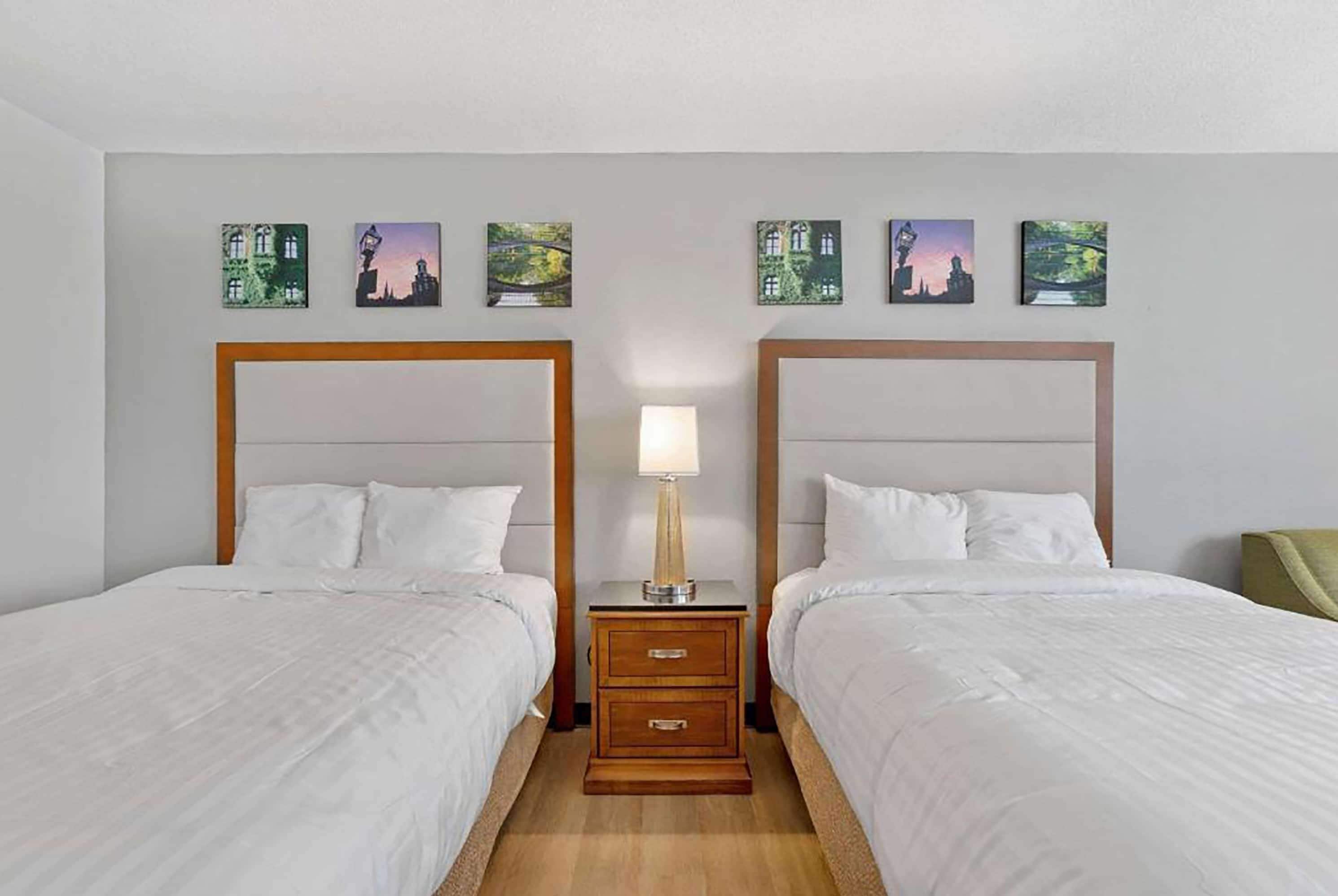 Habitación de hotel con dos camas, una mesita de noche con lámpara y cuatro cuadros de paisajes en la pared.