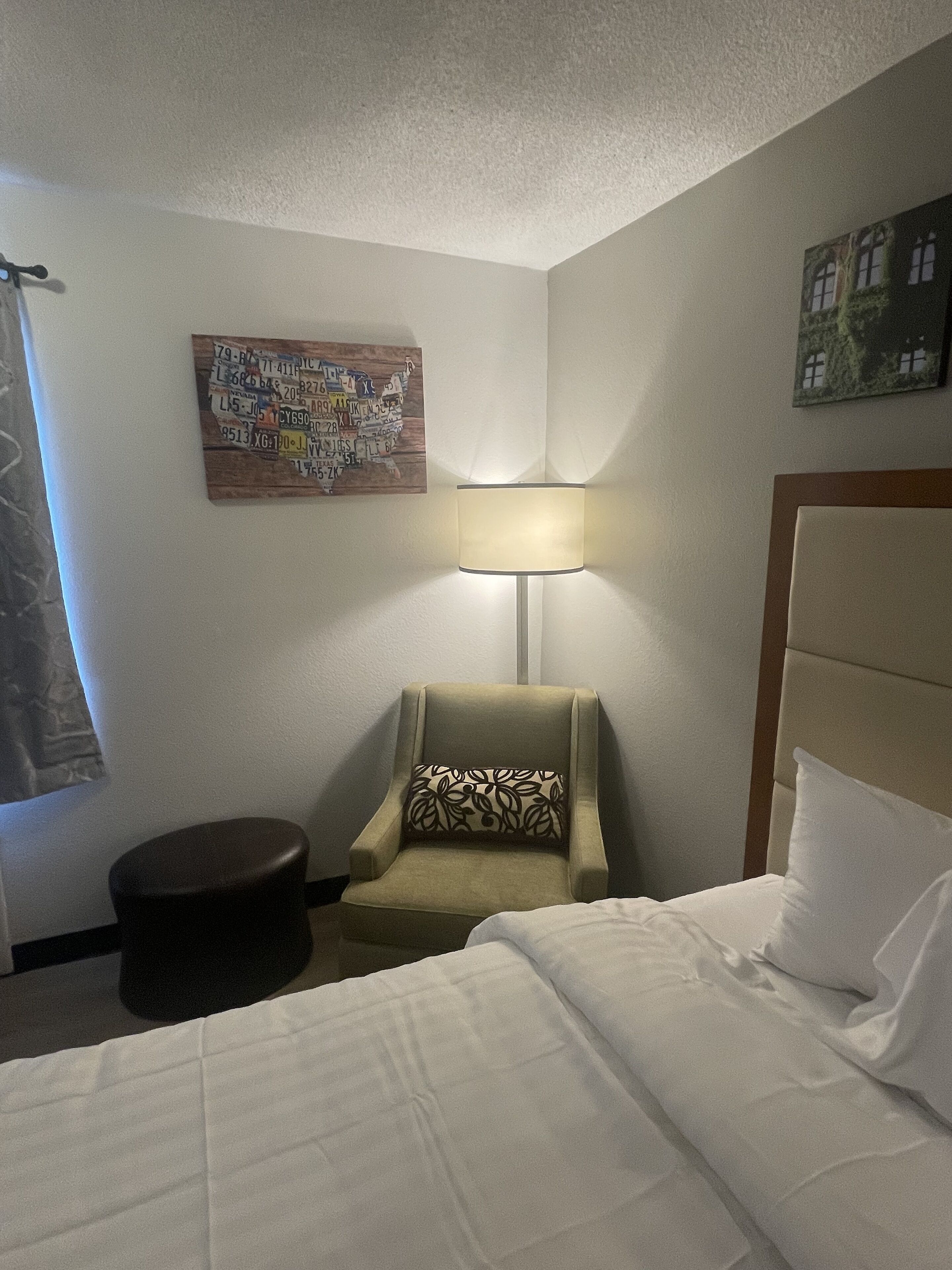 A hotel room with a bed, a chair with a patterned cushion, a wall-mounted lamp, and a framed picture.