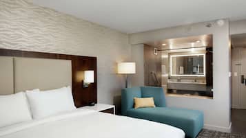 A hotel room with a large bed, a seating area with a blue chair, a bedside table with a lamp, and a bathroom with a mirror and shower.