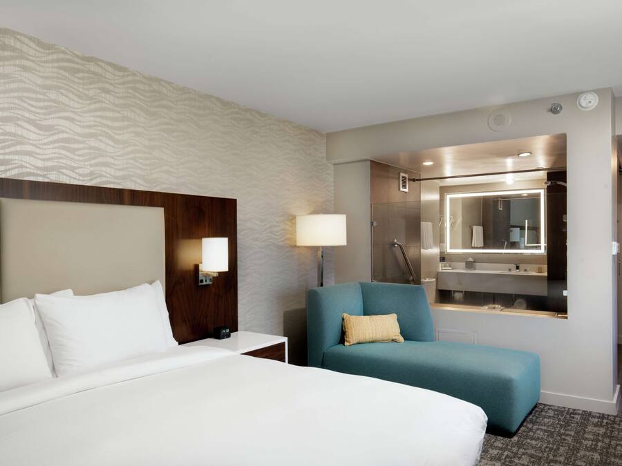 A hotel room with a large bed, a seating area with a blue chair, a bedside table with a lamp, and a bathroom with a mirror and shower.