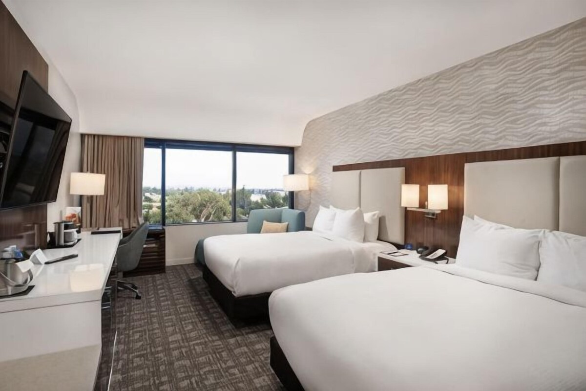 A hotel room with two beds, a desk, a TV, and a large window.