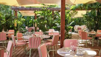 An outdoor dining area with round tables and chairs, surrounded by lush greenery.