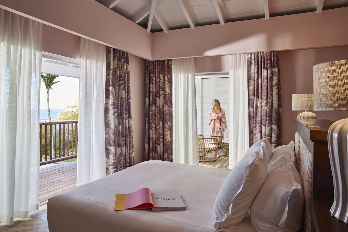 A hotel room with a large bed, a view of the balcony, and a person in a pink dress.