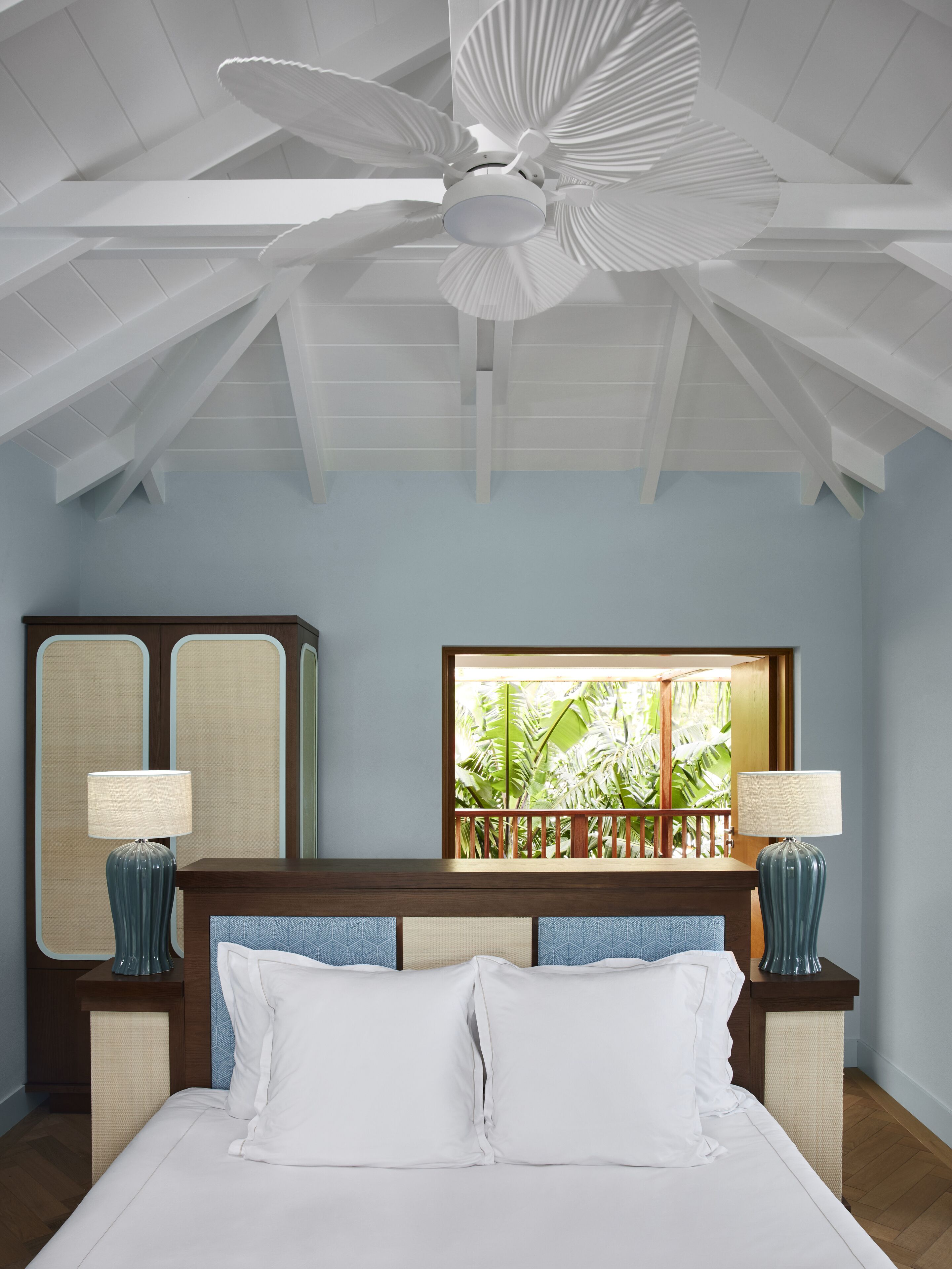 A bedroom with a bed, two bedside tables with lamps, a ceiling fan, a large window with a view of greenery, and a wooden headboard.