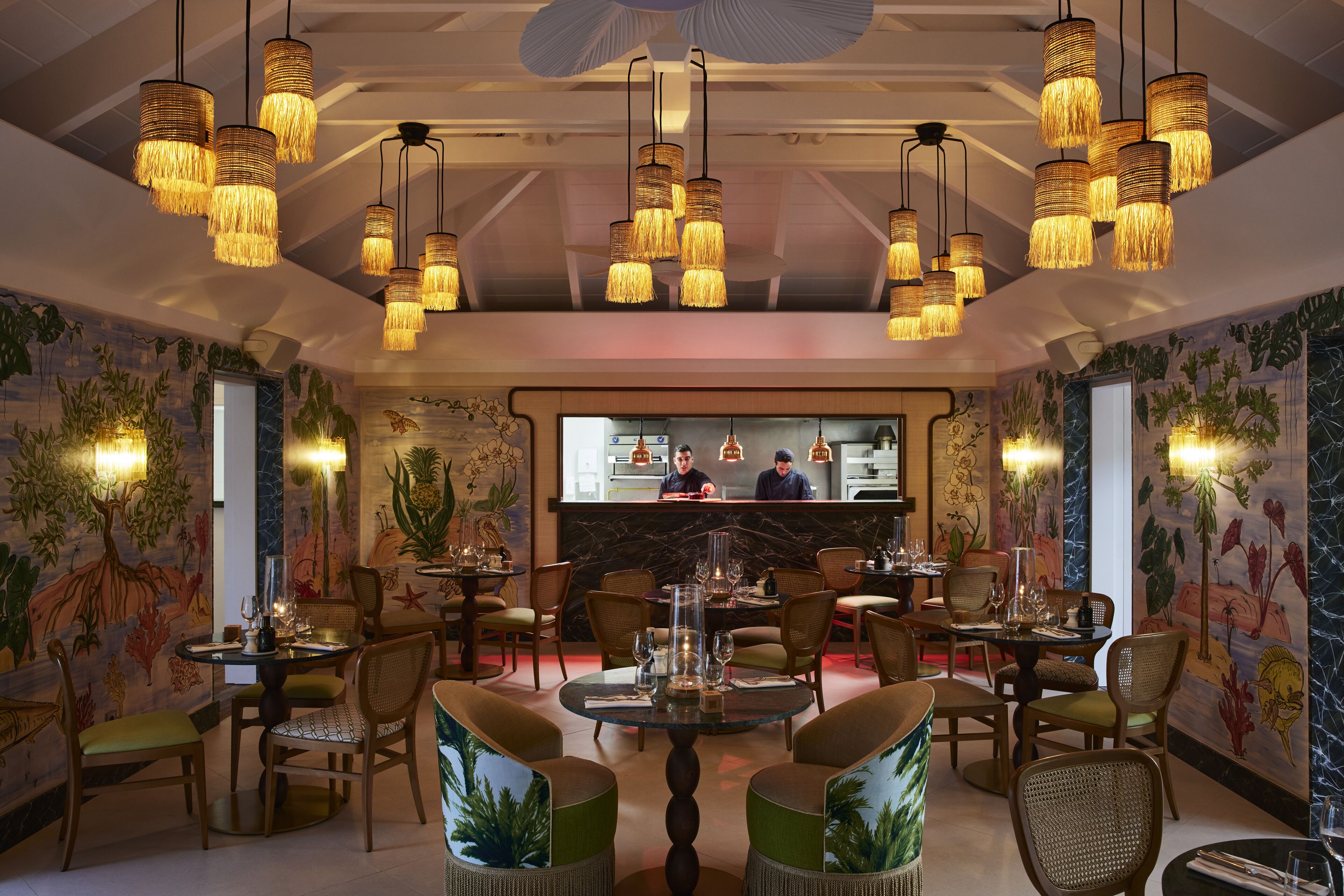 A restaurant with multiple dining tables and chairs, hanging wicker chandeliers, and a bar area with two staff members.