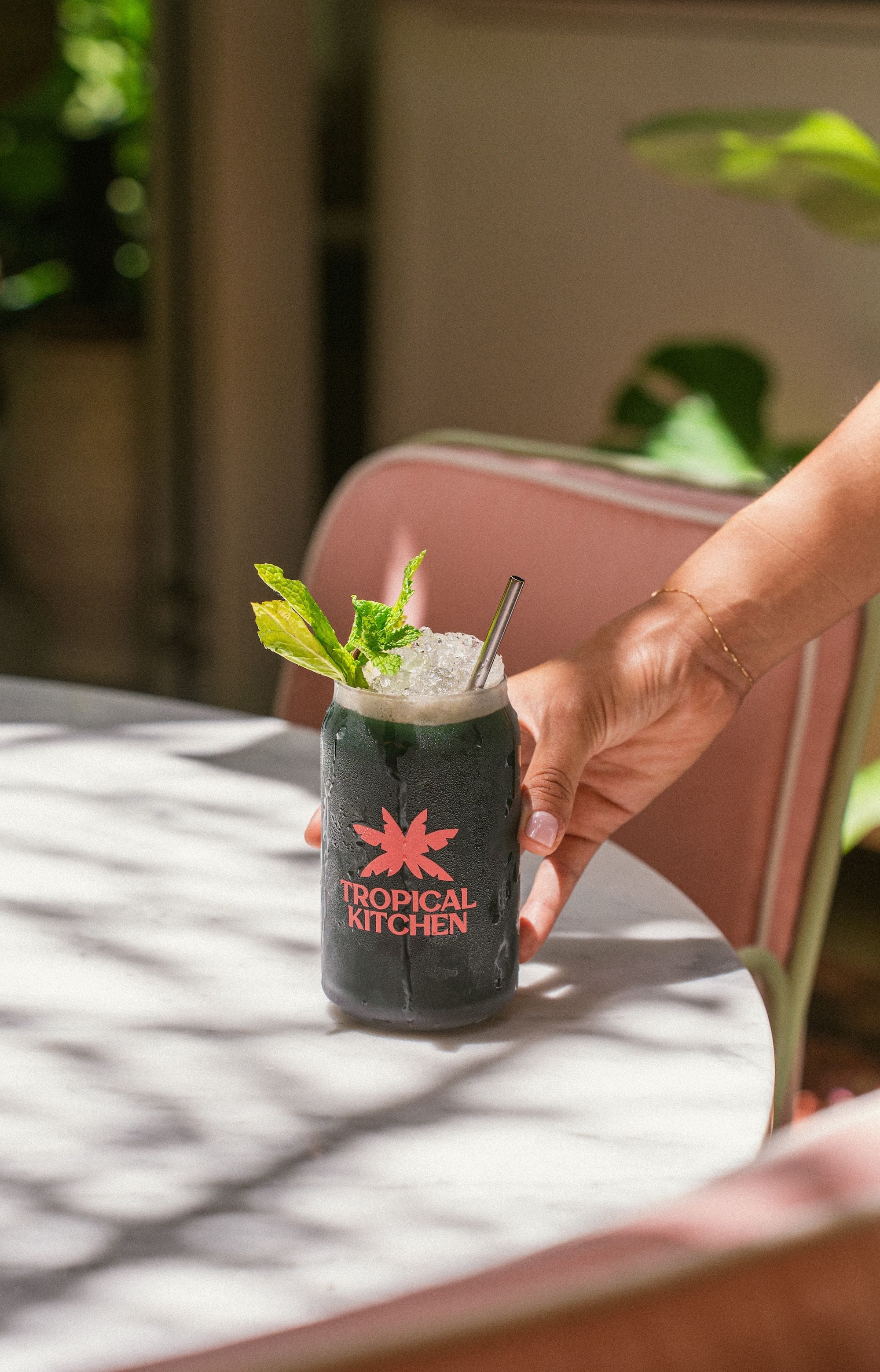A hand holding a glass with a straw, labeled 'TROPICAL KITCHEN,' on a table with a white surface and a pink chair in the background.