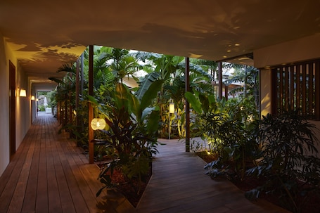 A wooden pathway flanked by lush tropical plants and ambient lighting, leading to a building entrance.