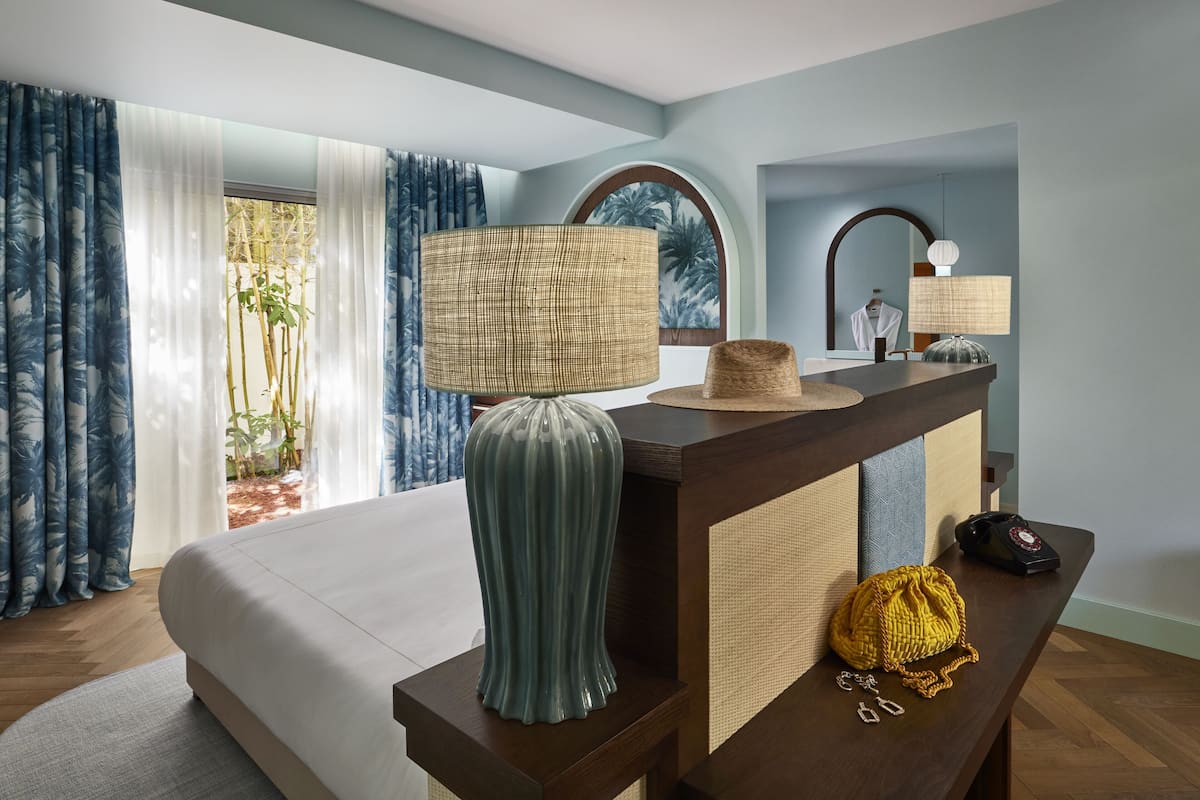 A hotel room with a bed, a bedside table with a lamp, a decorative vase, and a small round table with a hat and a phone.