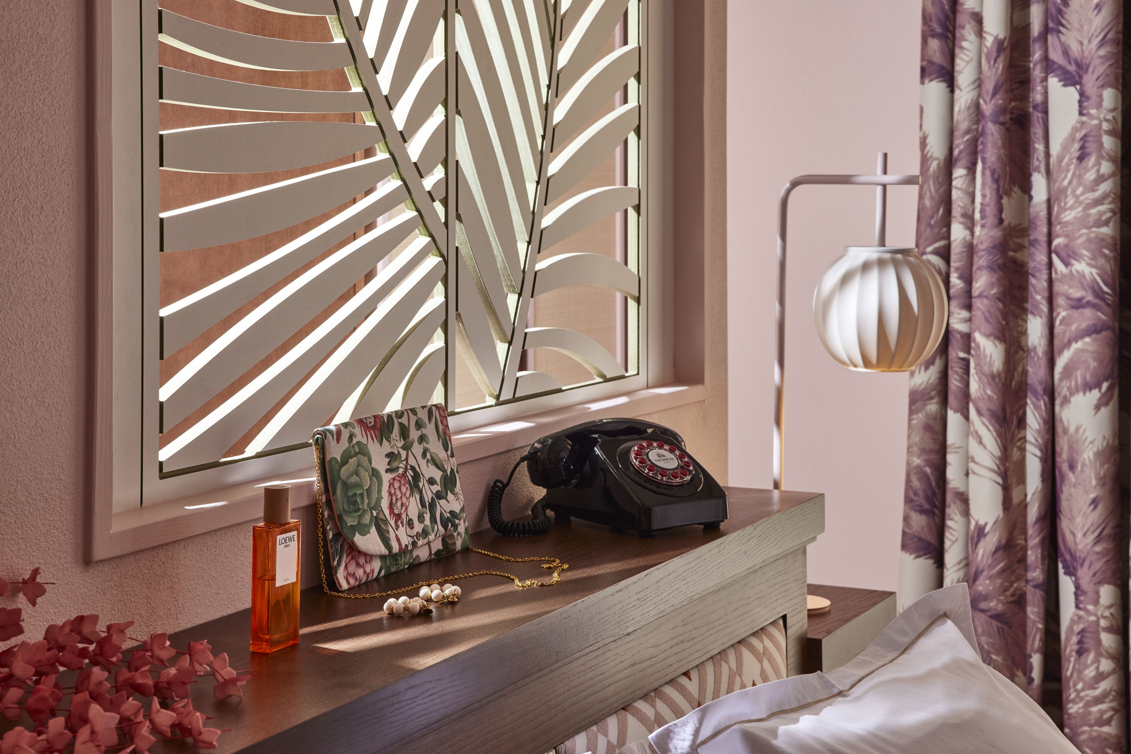A neatly arranged hotel room with a wooden bedside table, a vintage rotary phone, a floral-patterned bag, a perfume bottle, and a decorative wall panel.