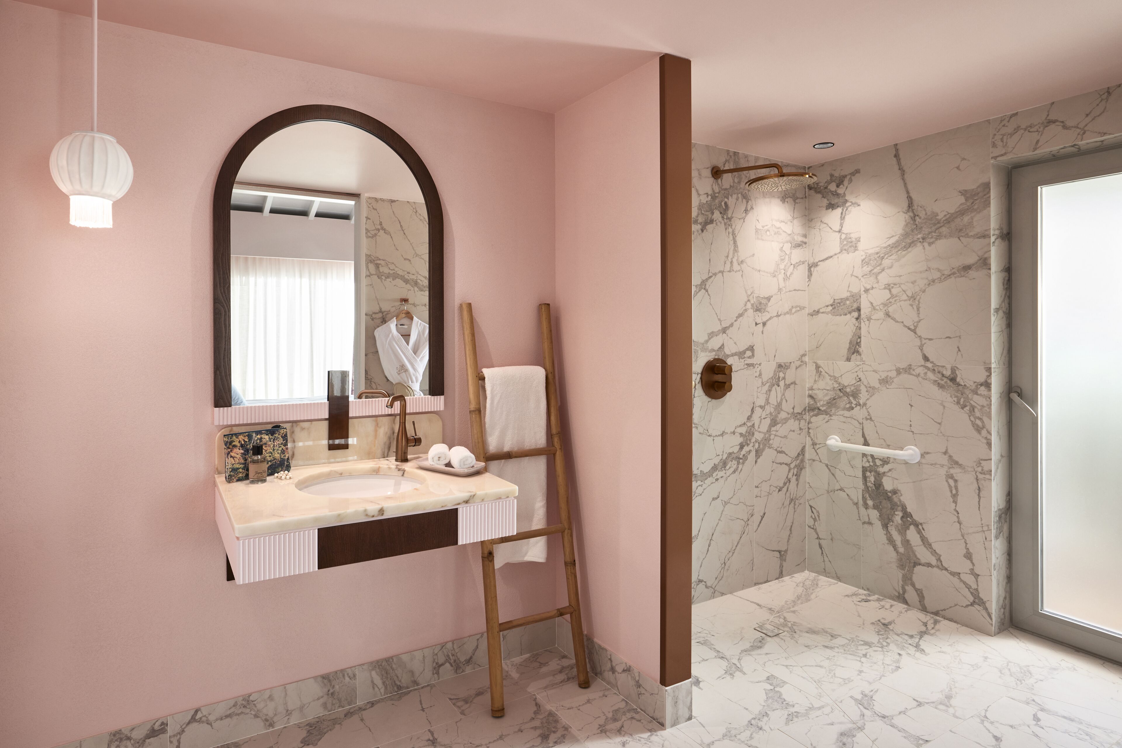A bathroom with a pink wall, marble flooring, a sink with a mirror, and a shower area.