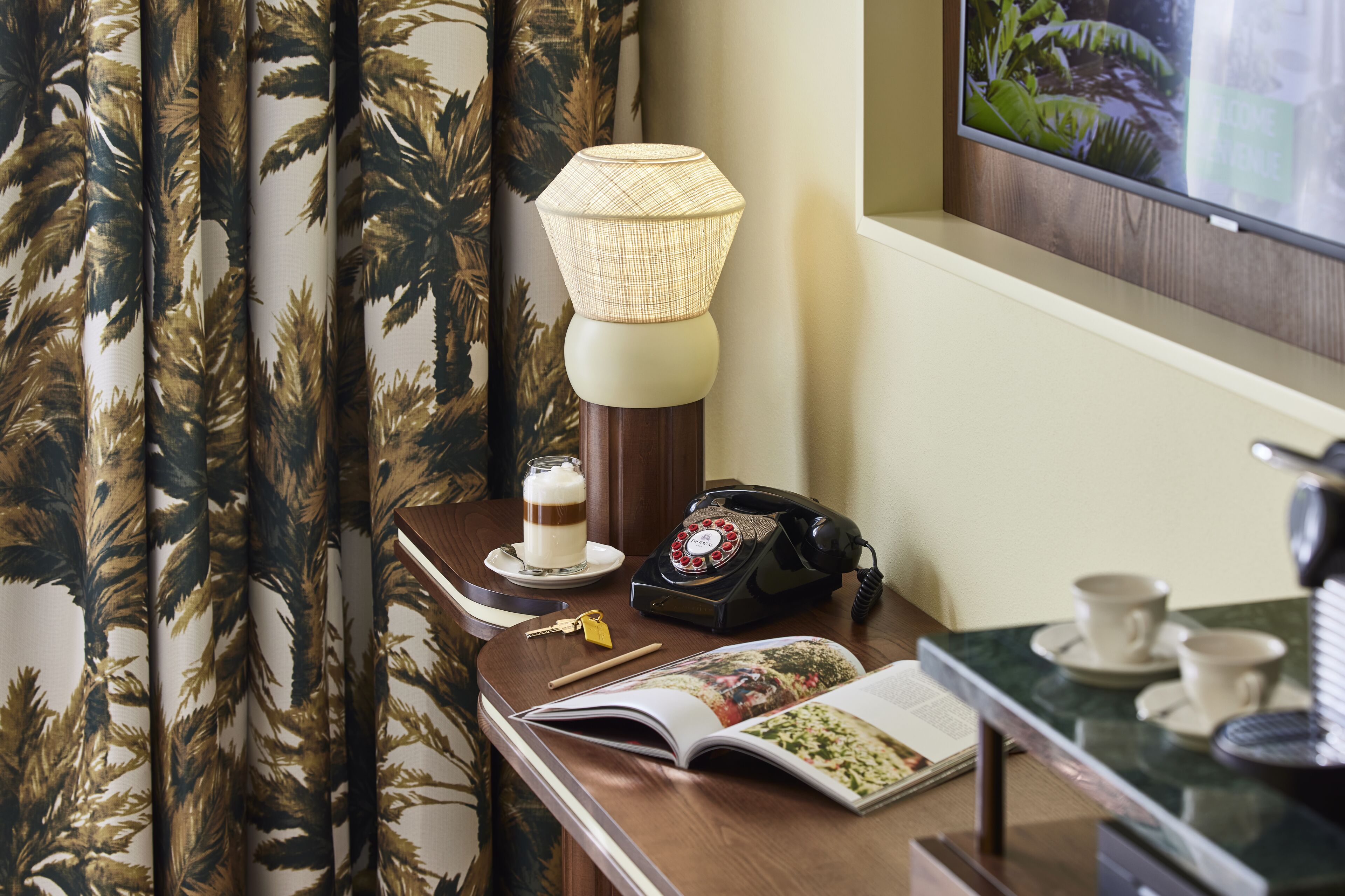Una camera d'albergo con una scrivania in legno, una lampada, un telefono, un libro e una tazza su un piattino.