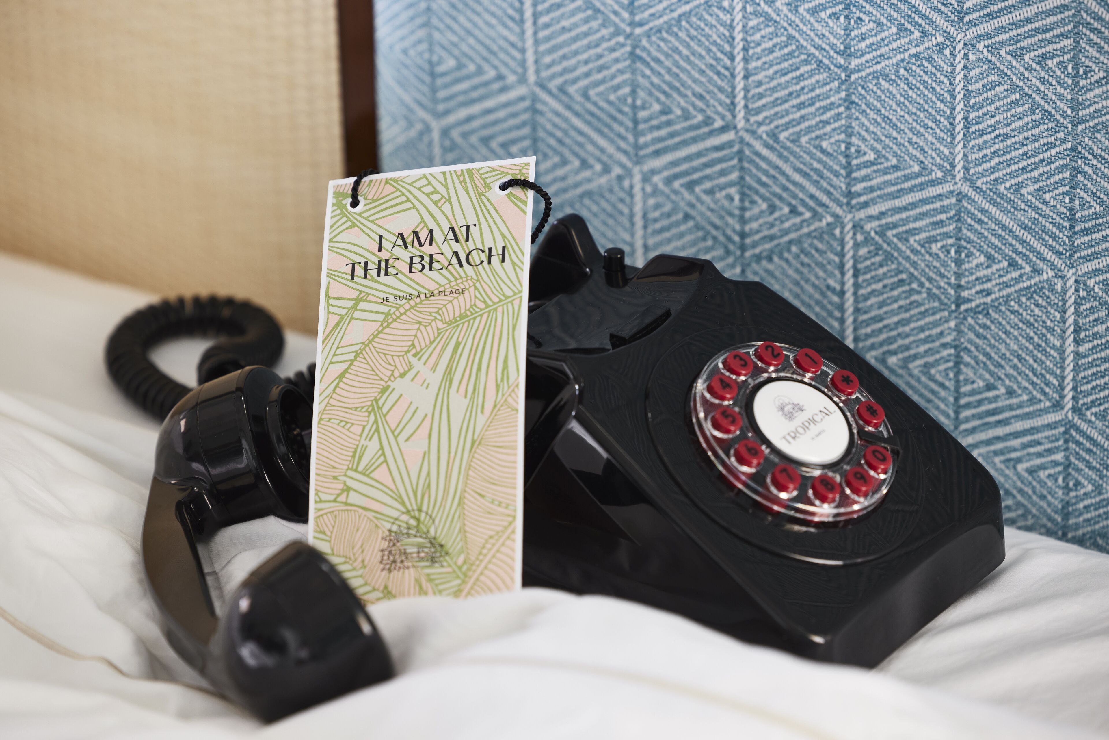 A vintage rotary phone, a postcard, and a black telephone handset on a bed with patterned wallpaper.