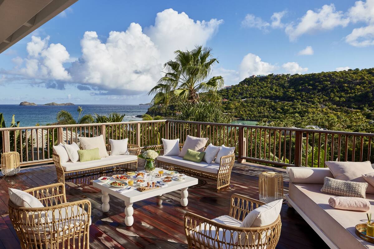 A deck with wicker furniture, a dining table with food, and a view of the ocean and hills.