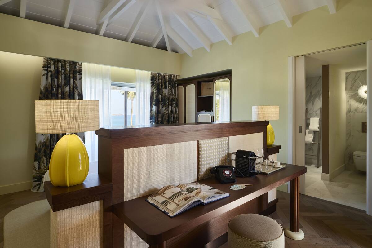 A hotel room with a wooden headboard, a bedside table with a lamp, a mirror, and a view of the bathroom.