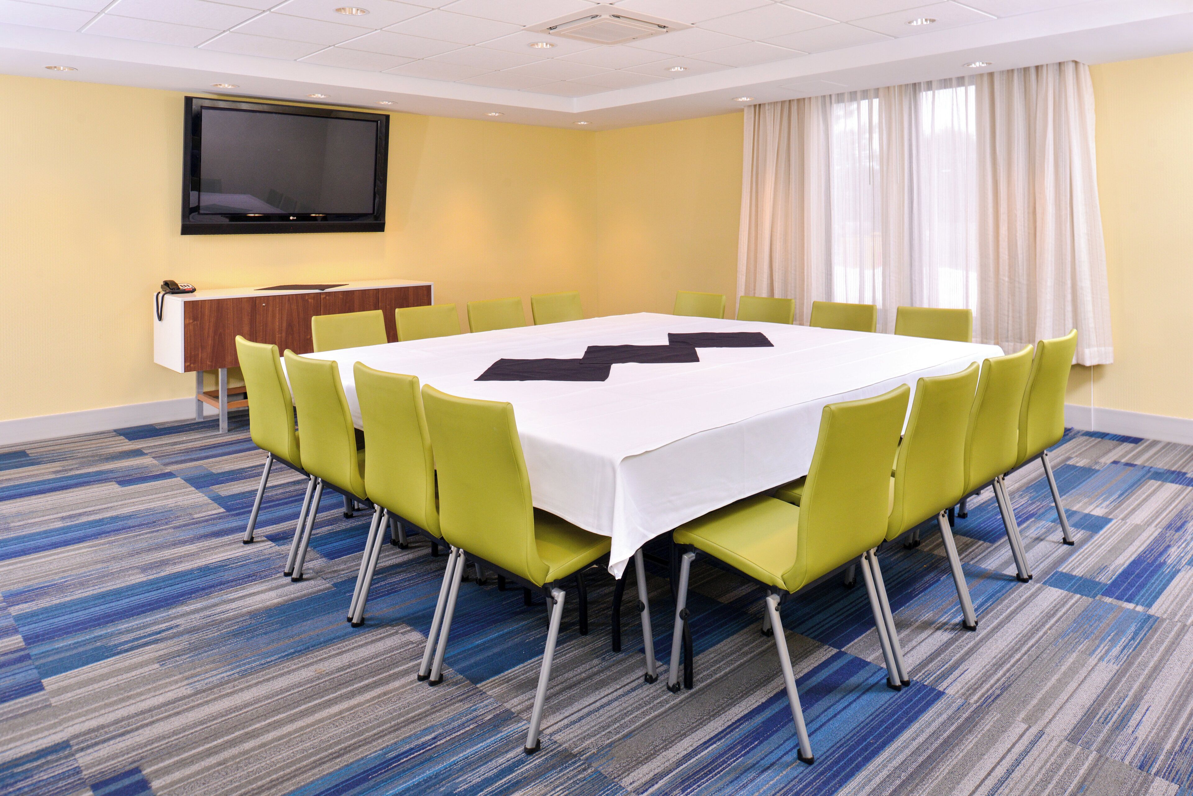 A conference room with a large table, green chairs, a flat-screen TV, and a telephone on a desk.