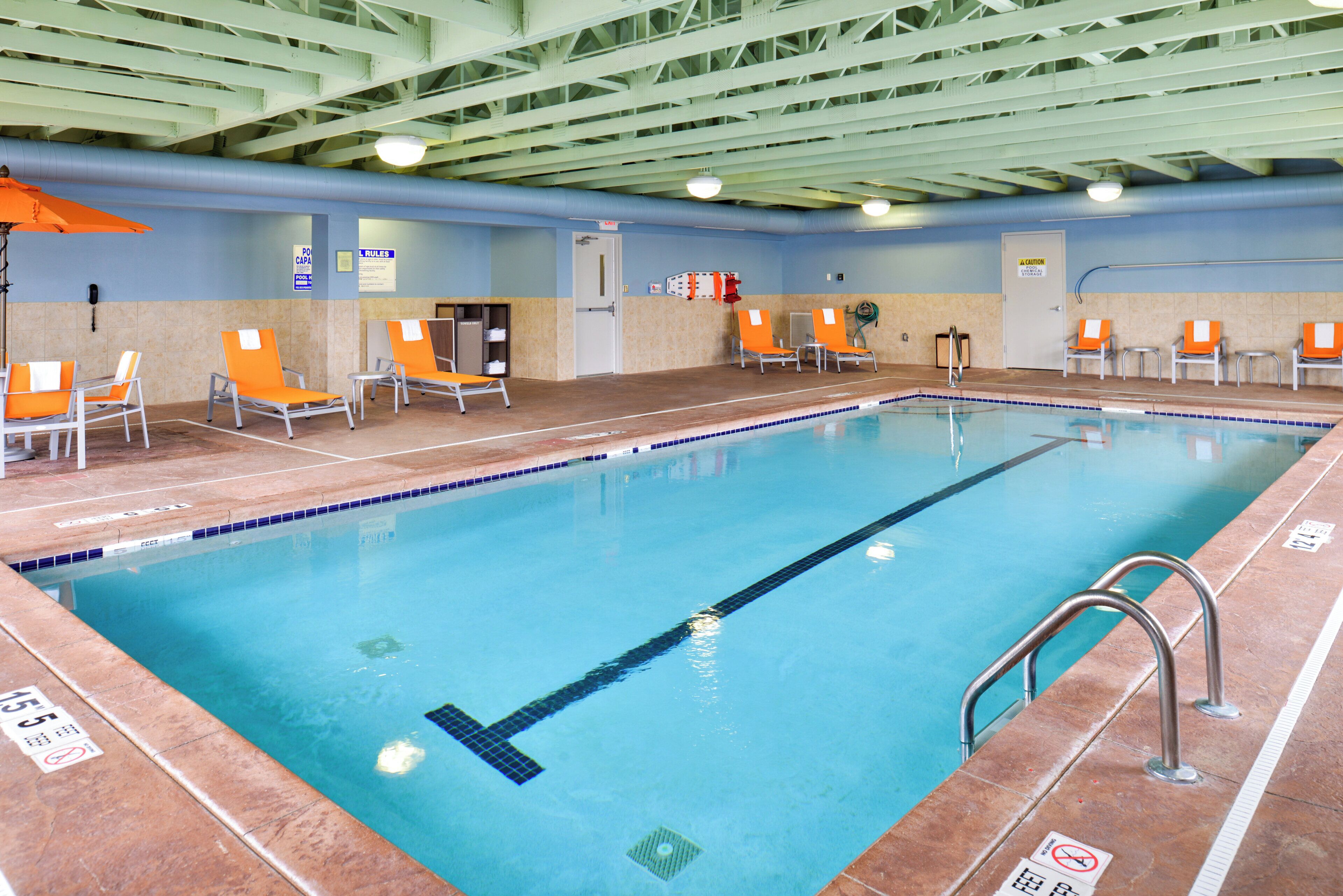 An indoor swimming pool with clear lane markings, a lifeguard chair, and a poolside area with chairs and a towel rack.