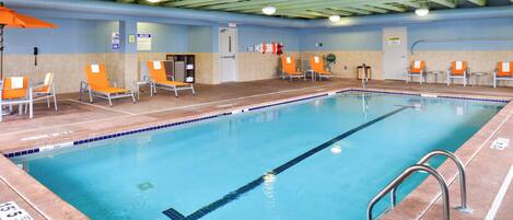An indoor swimming pool with clear lane markings, a lifeguard chair, and a poolside area with chairs and a towel rack.