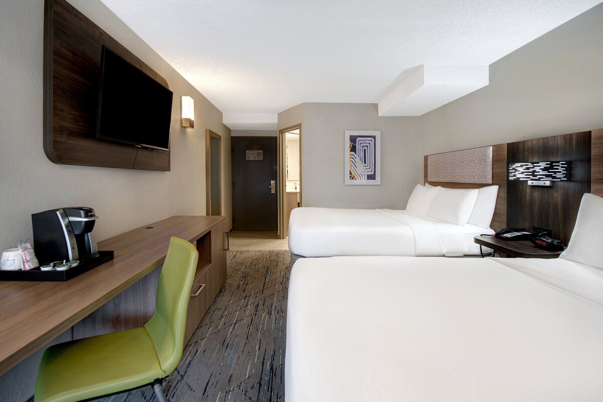 A hotel room with a bed, a desk with a coffee maker, a chair, a TV mounted on the wall, and a door leading to another room.