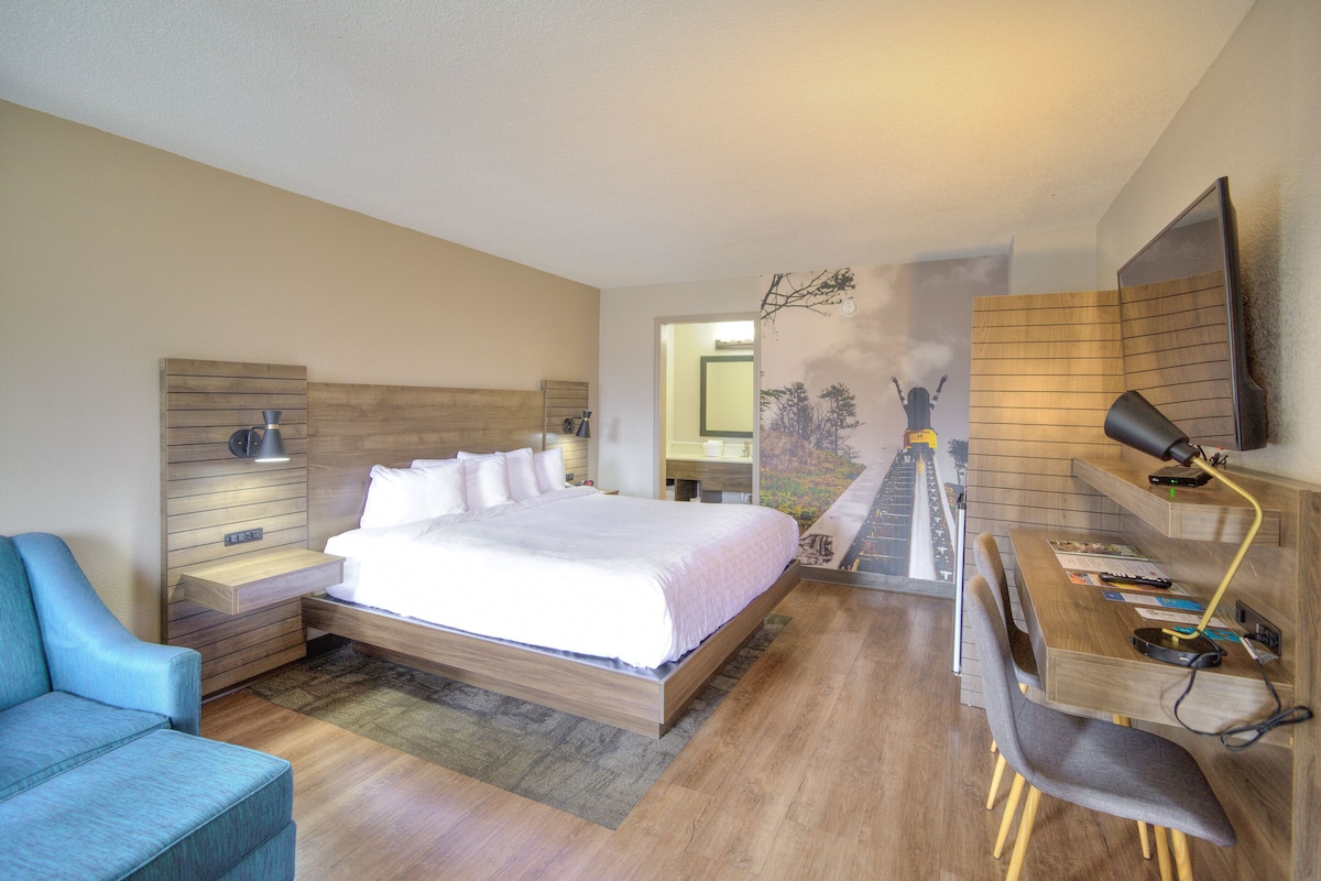 Ein modernes Hotelzimmer mit einem großen Bett, einem Schreibtisch mit Stuhl, einem Fernseher und einem an der Wand befestigten Regal.