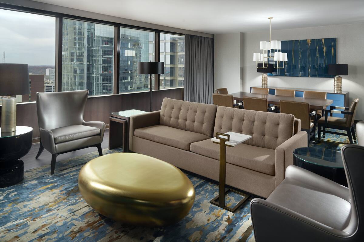 A modern hotel room with a large window, a sofa, a coffee table, and a dining area.