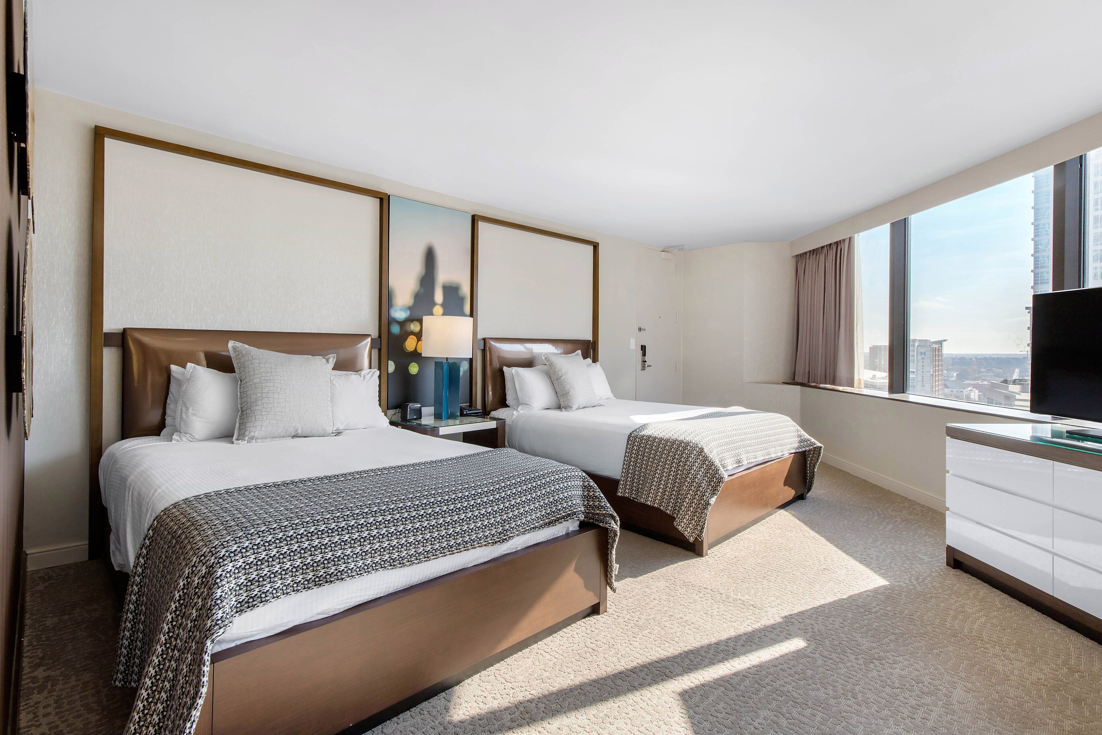 A hotel room with two beds, a TV, a desk, and a large window offering a city view.