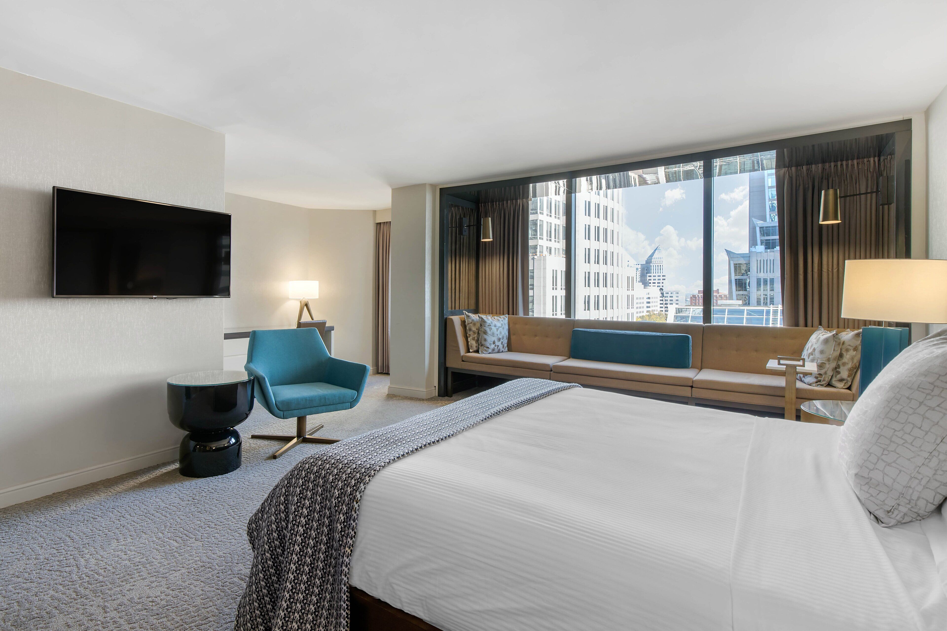 Ein Hotelzimmer mit einem großen Bett, einem blauen Sessel, einer Couch, einem Flachbildfernseher und einem Fenster mit Blick auf die Stadt.