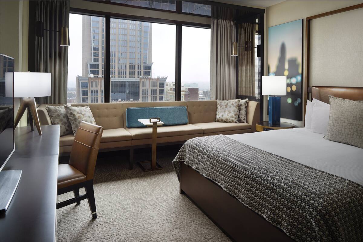 A hotel room with a large bed, a sofa, a desk, and a view of the cityscape through large windows.