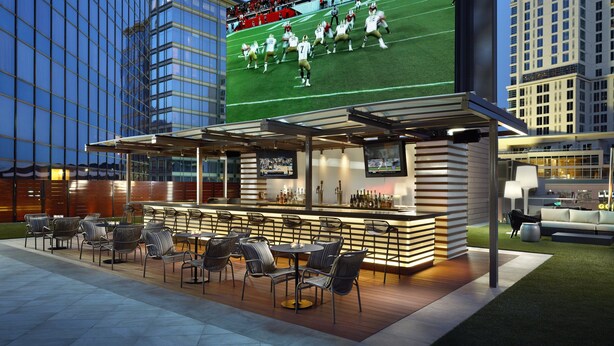 An outdoor bar area with seating and a large screen displaying a football game.