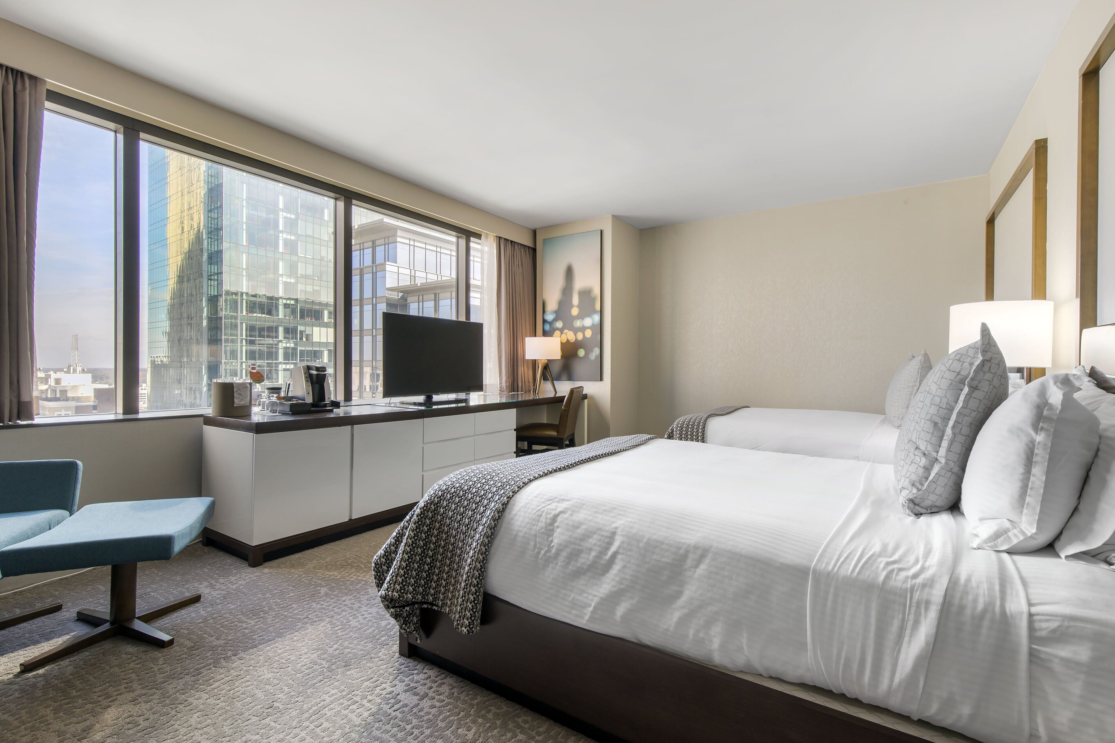 A hotel room with a large bed, a desk with a flat-screen TV, a chair, and a view of a cityscape through large windows.