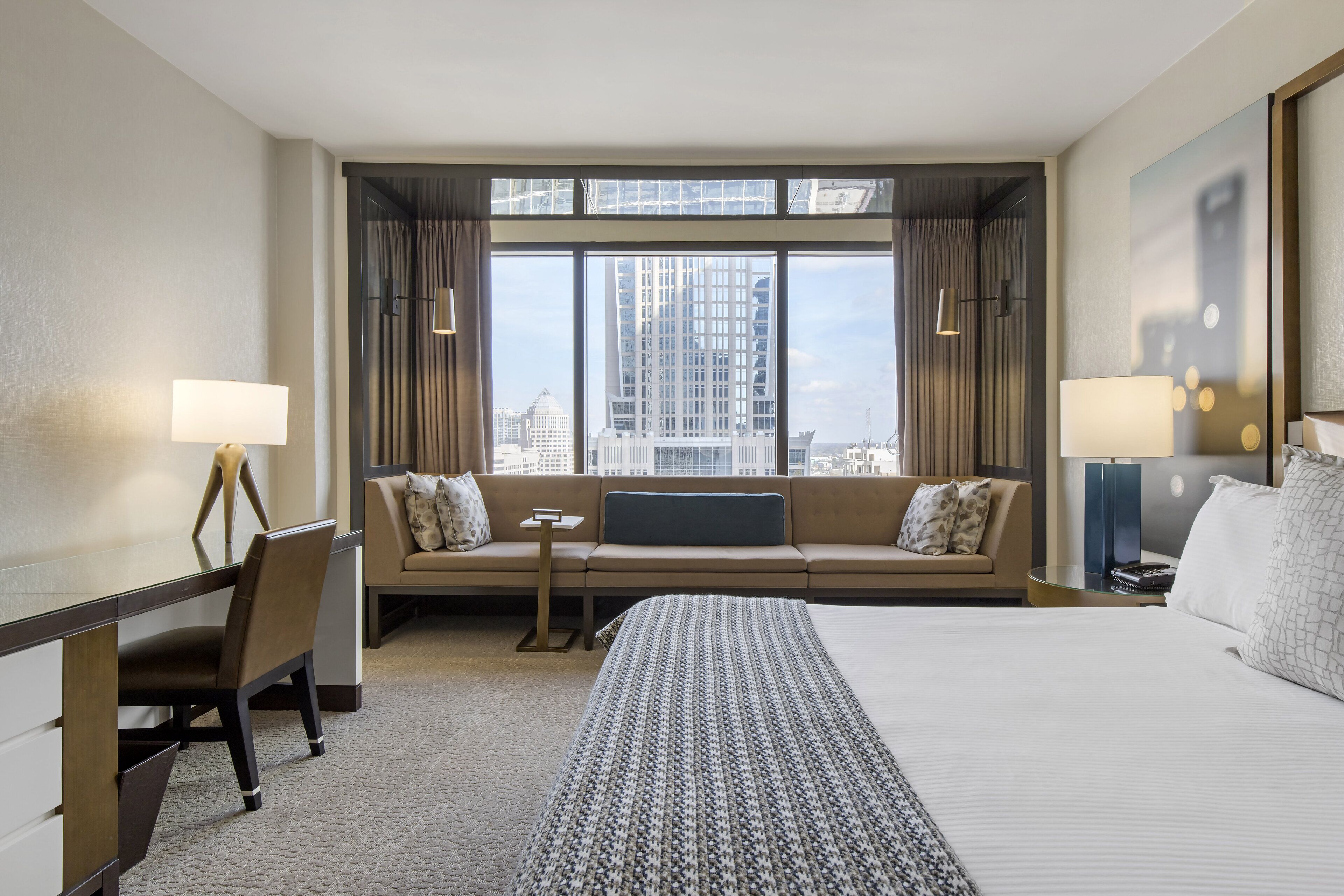 A hotel room with a large bed, a sofa, a desk, and a view of the city through large windows.