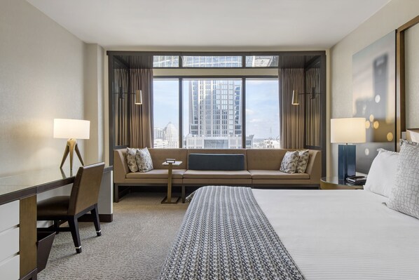 A hotel room with a large bed, a sofa, a desk, and a view of the city through large windows.