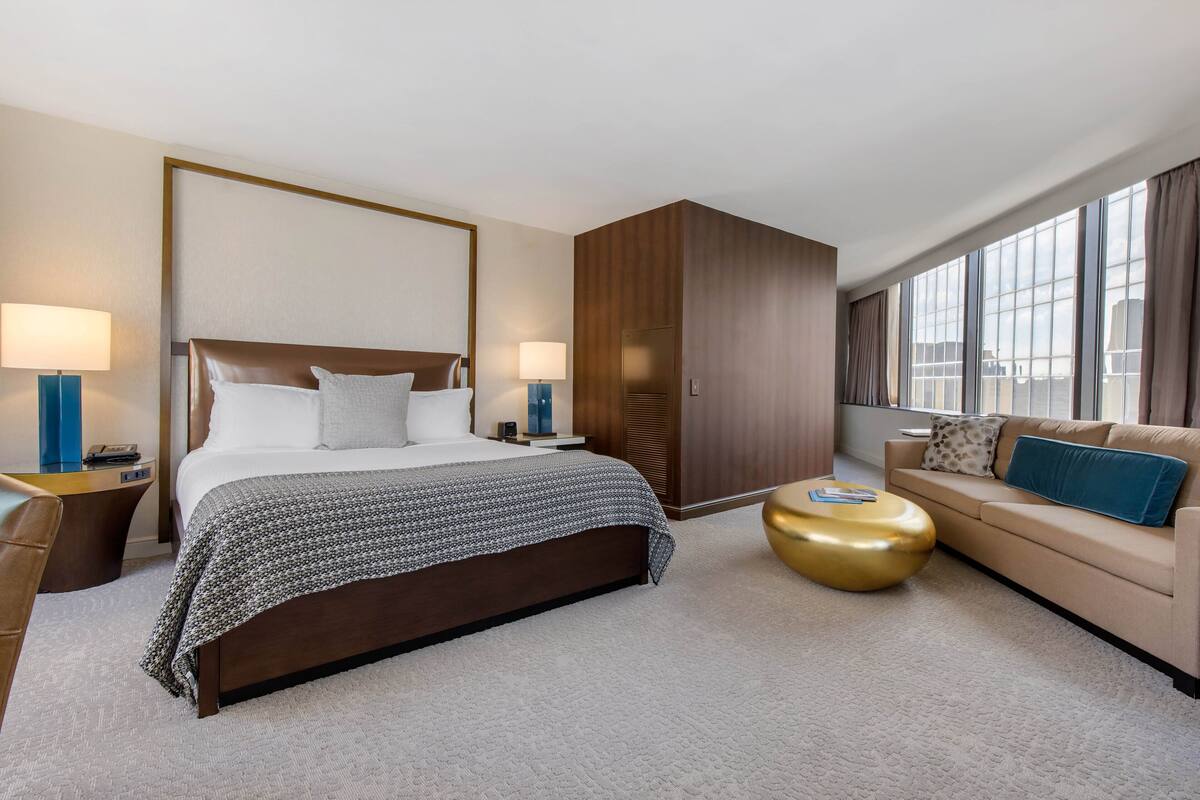 A hotel room with a large bed, a sofa, a gold coffee table, and a large window with curtains.