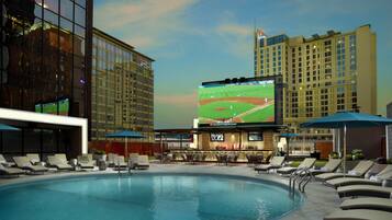 A swimming pool with lounge chairs and umbrellas, a bar area, and a large screen displaying a baseball game.